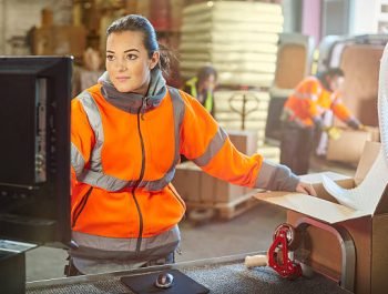 a female warehouse packer checks the order on the computer whilst packing boxes in the despatch area of a warehouse. In the background a male and female co-worker are seen loading the back of a van .