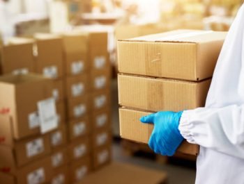 Close up view of a dedicated worker carrying a stack of duck taped brown boxes in factory storage room while wearing sterile cloths and rubber gloves.